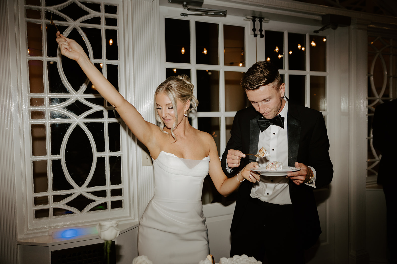 Bride raises her fork in celebration while the groom eats a slice of cake during the reception.